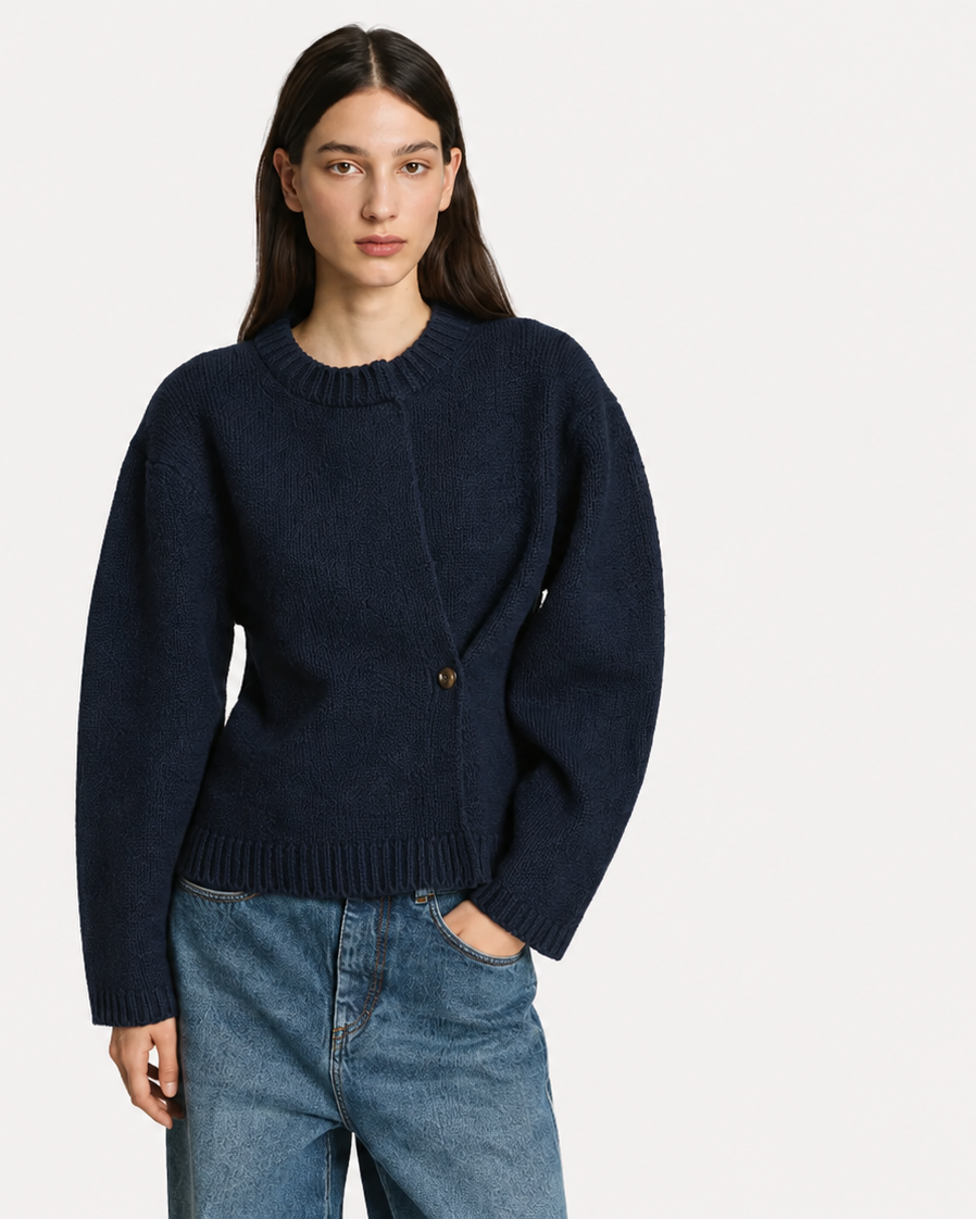 A woman modeling a dark navy blue knit sweater with a unique asymmetrical button closure, paired with blue jeans, against a plain light gray background.