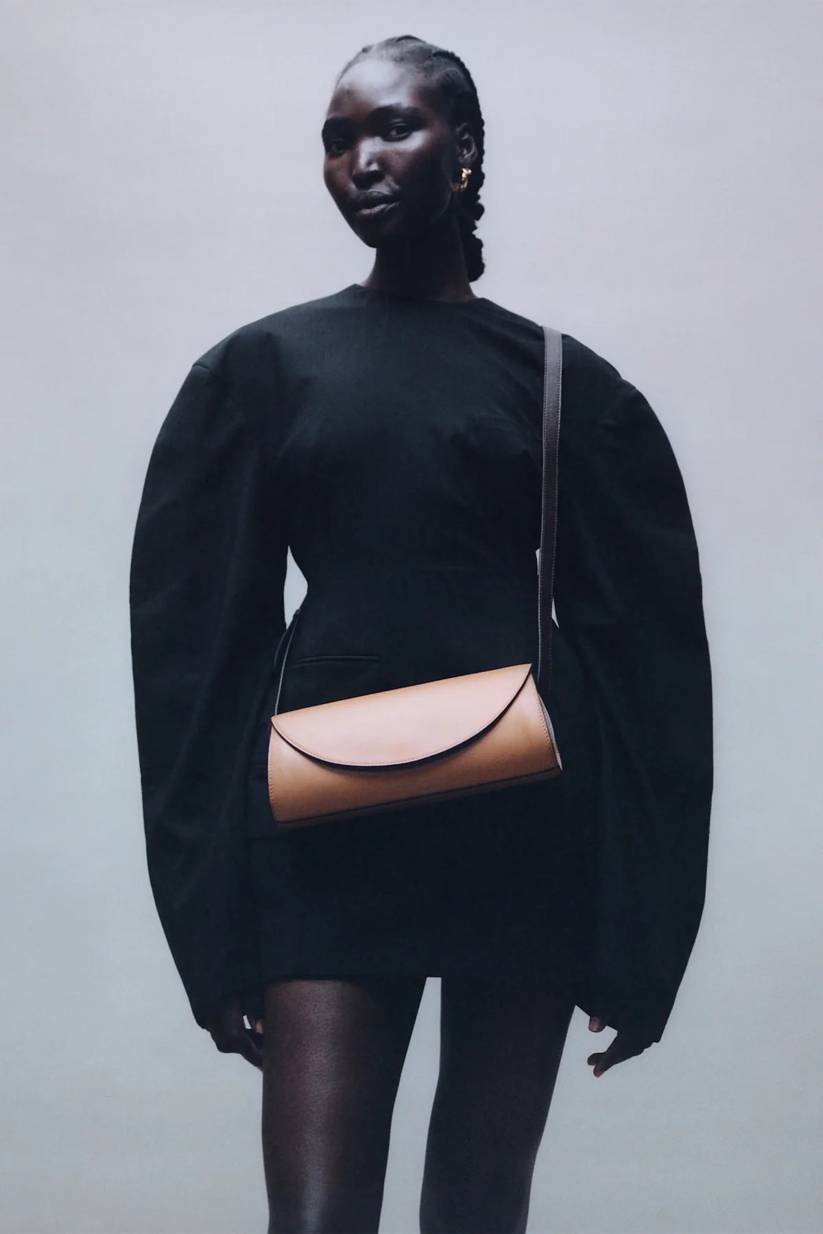 Person wearing a black outfit with a tan leather bag on a plain background
