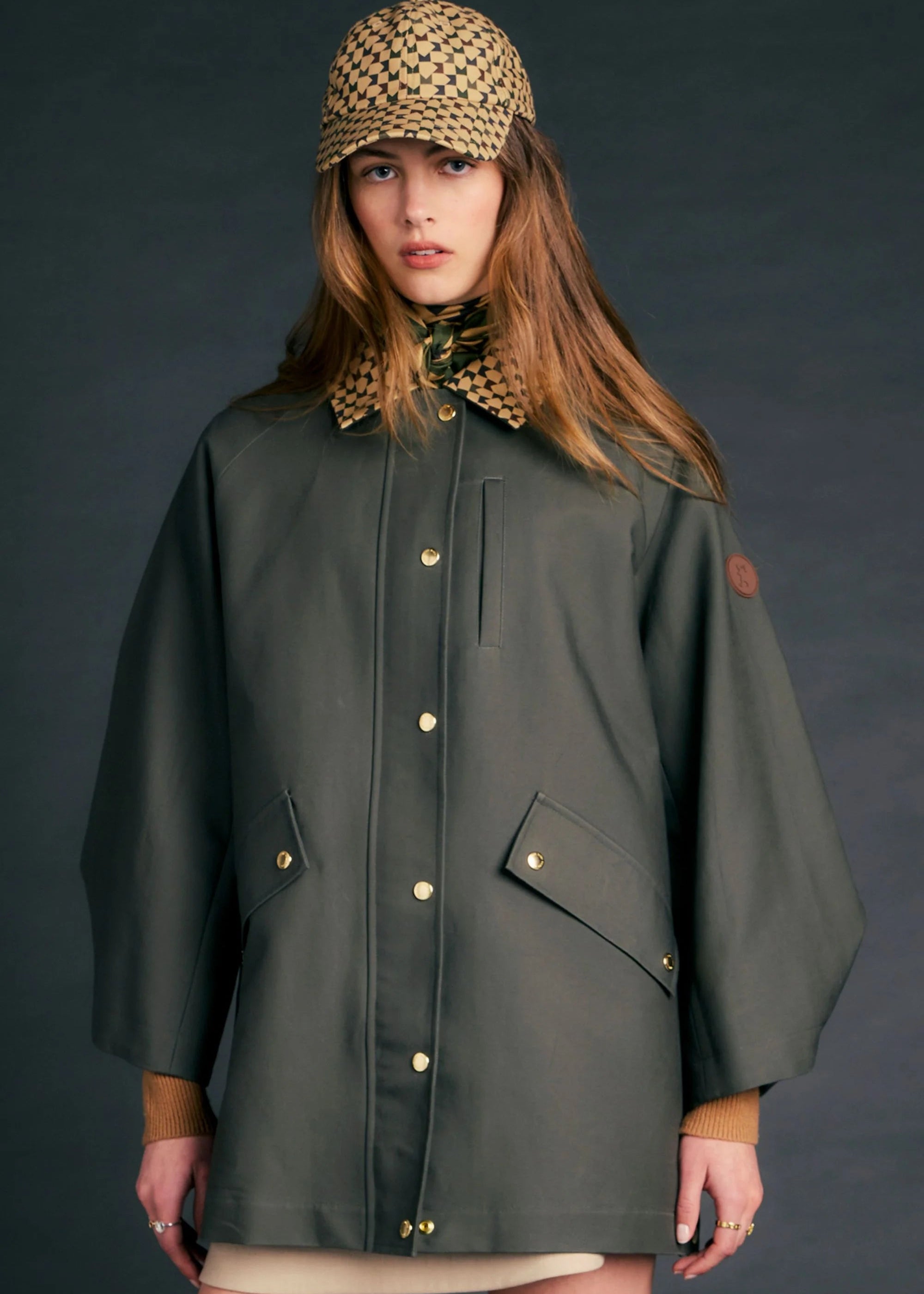 A model wearing a dark olive green utility jacket with gold-tone snap buttons, a patterned knit cap, and a beige skirt, posed against a dark gray studio background.
