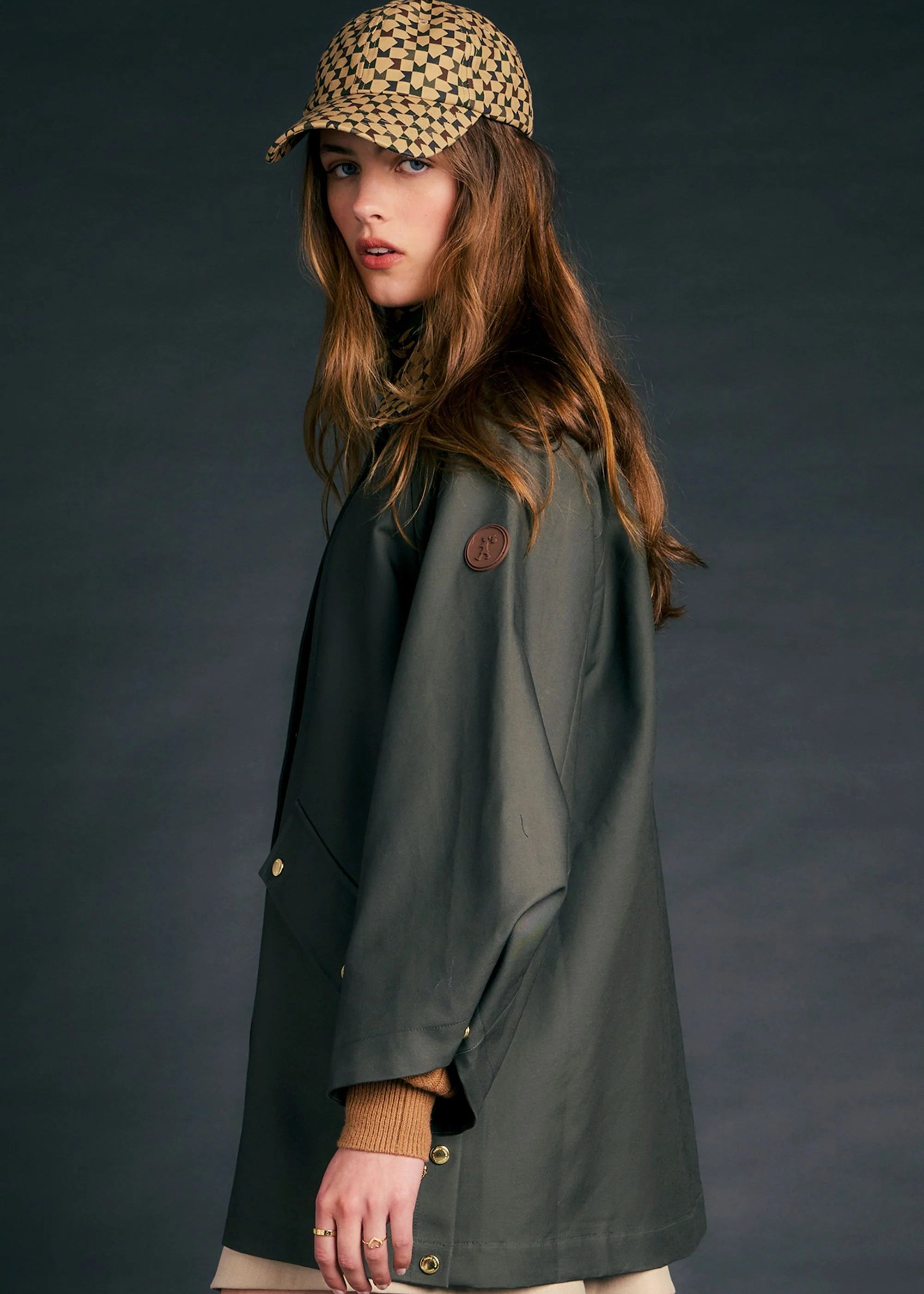 A woman wearing a dark olive green raincoat, a beige and black checkered cap, and a beige skirt, photographed against a dark gray studio background.