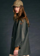 A woman wearing a dark olive green raincoat, a beige and black checkered cap, and a beige skirt, photographed against a dark gray studio background.