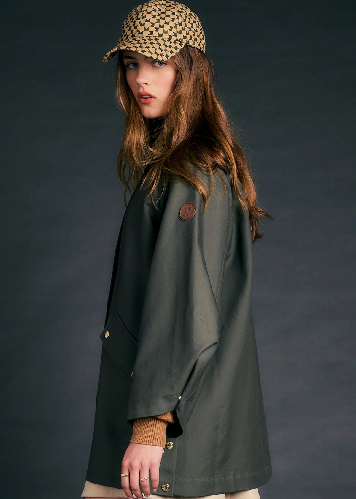 A woman wearing a dark olive green raincoat, a beige and black checkered cap, and a beige skirt, photographed against a dark gray studio background.