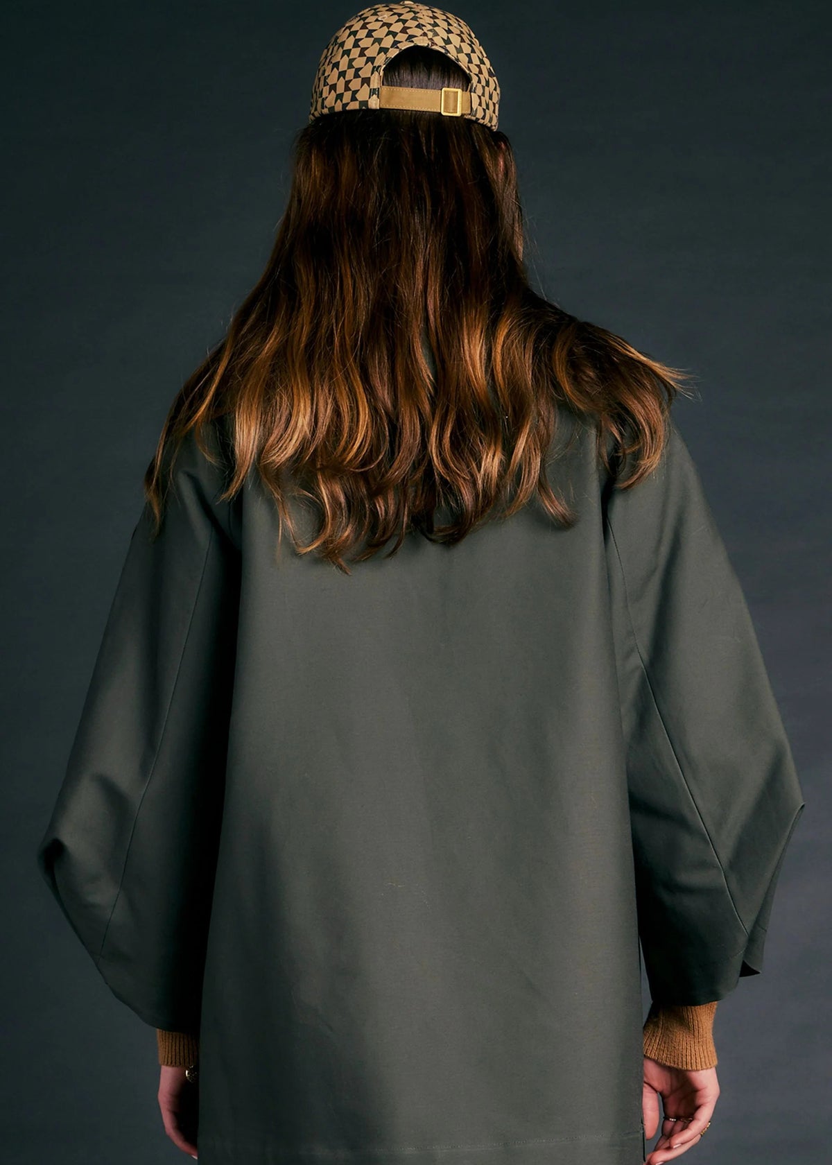 A person shown from the back wearing a dark olive green oversized coat with wide sleeves, a patterned beige and black baseball cap, and light-colored pants, set against a dark gray studio background.