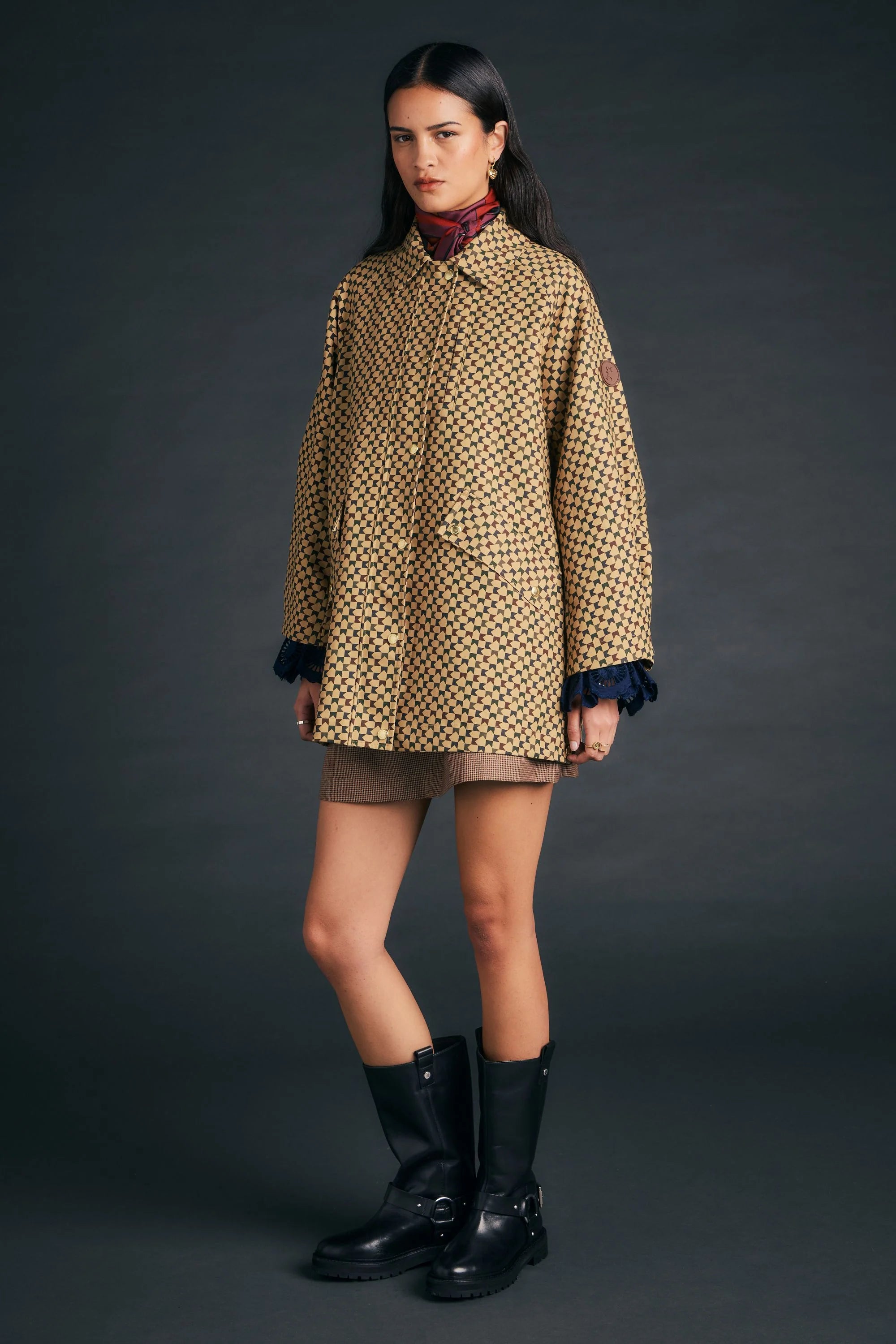 A model stands against a dark gray studio background wearing a beige and black patterned coat with dark blue ruffled cuffs, a brown mini skirt, and black boots.