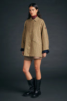 A model stands against a dark gray studio background wearing a beige and black patterned coat with dark blue ruffled cuffs, a brown mini skirt, and black boots.