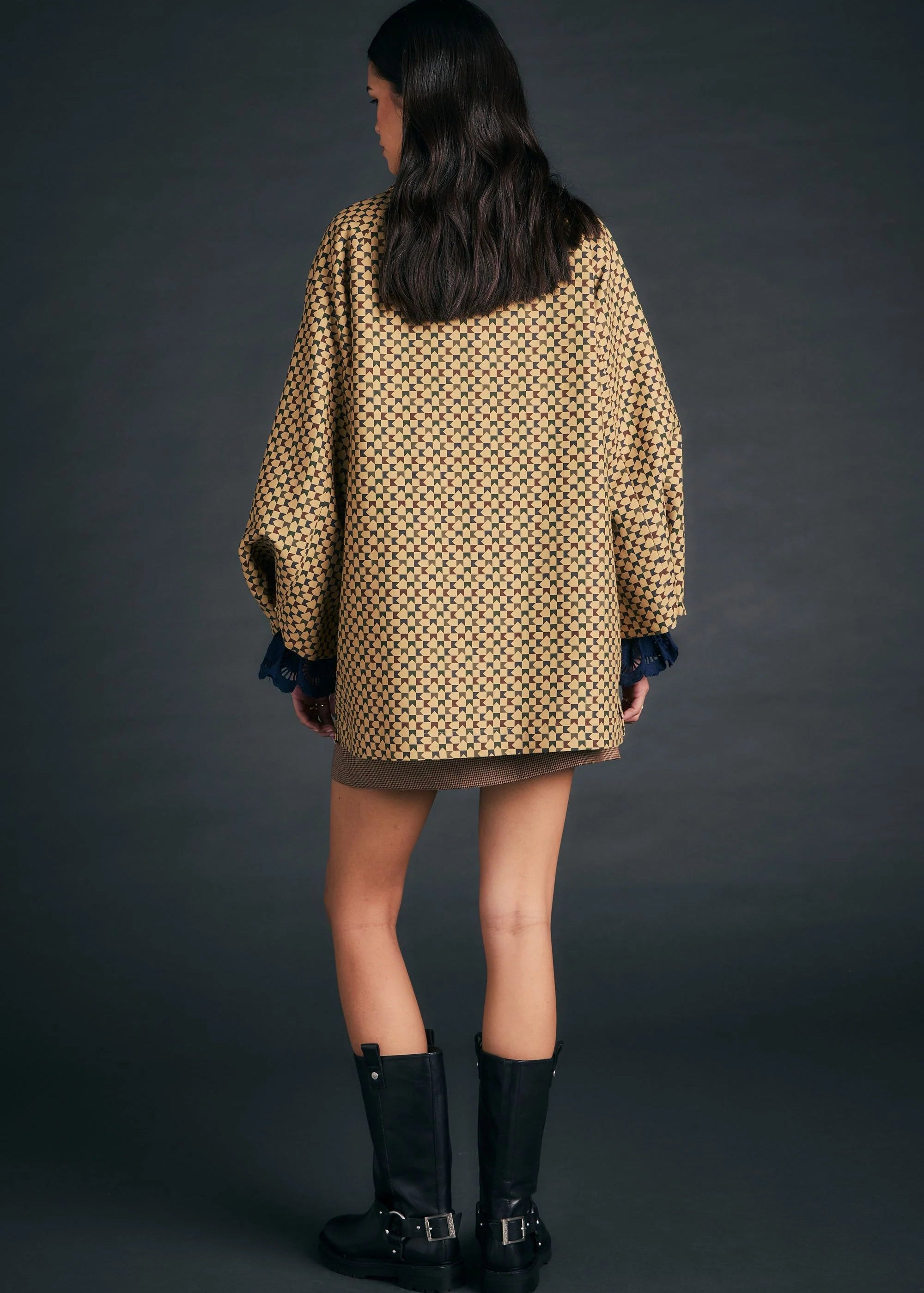 A model shown from the back wearing an oversized beige and black patterned coat with dark blue cuffs, a brown mini skirt, and black leather boots against a dark gray studio background.