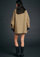 A model shown from the back wearing an oversized beige and black patterned coat with dark blue cuffs, a brown mini skirt, and black leather boots against a dark gray studio background.