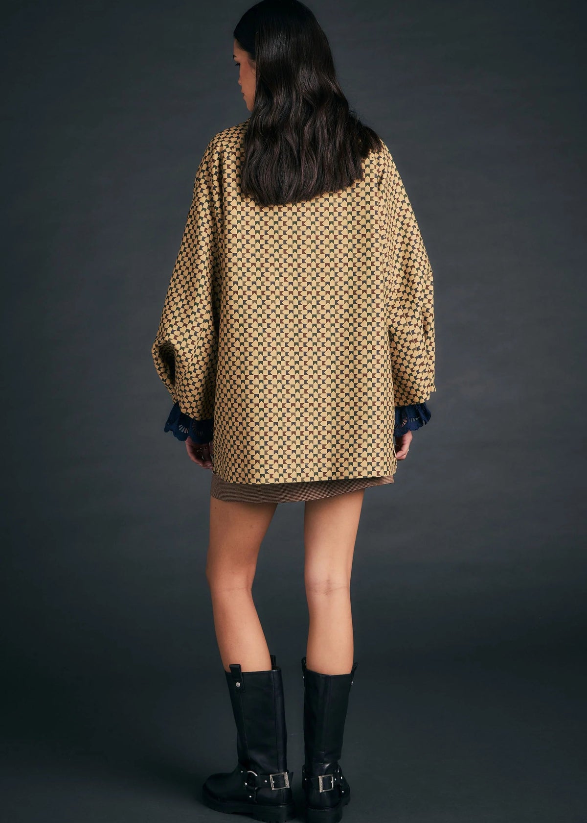 A model shown from the back wearing an oversized beige and black patterned coat with dark blue cuffs, a brown mini skirt, and black leather boots against a dark gray studio background.