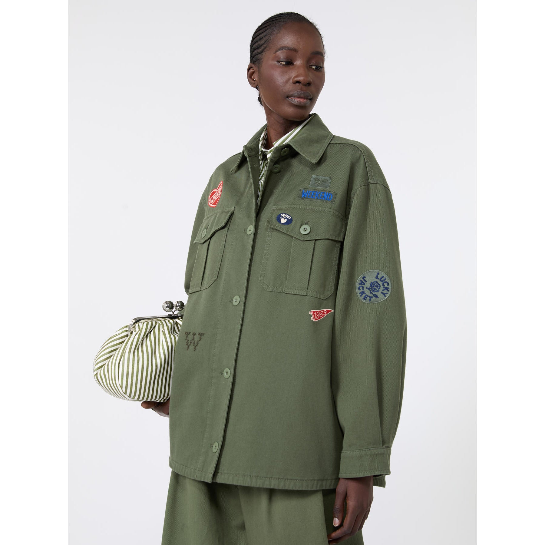 Person wearing a green jacket with patches and holding a striped bag on a white background