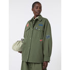 Person wearing a green jacket with patches and holding a striped bag on a white background