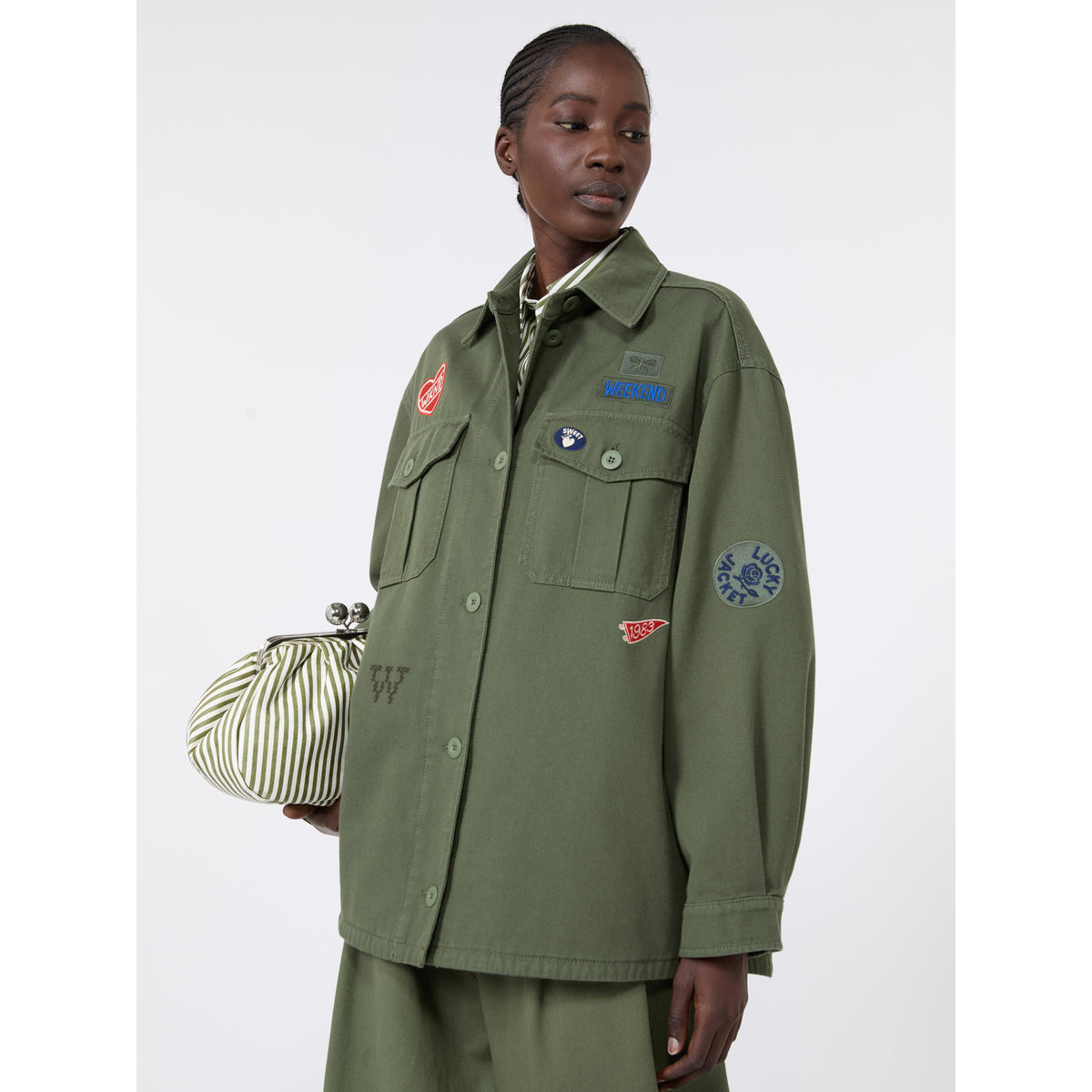 Person wearing a green jacket with patches and holding a striped bag on a white background