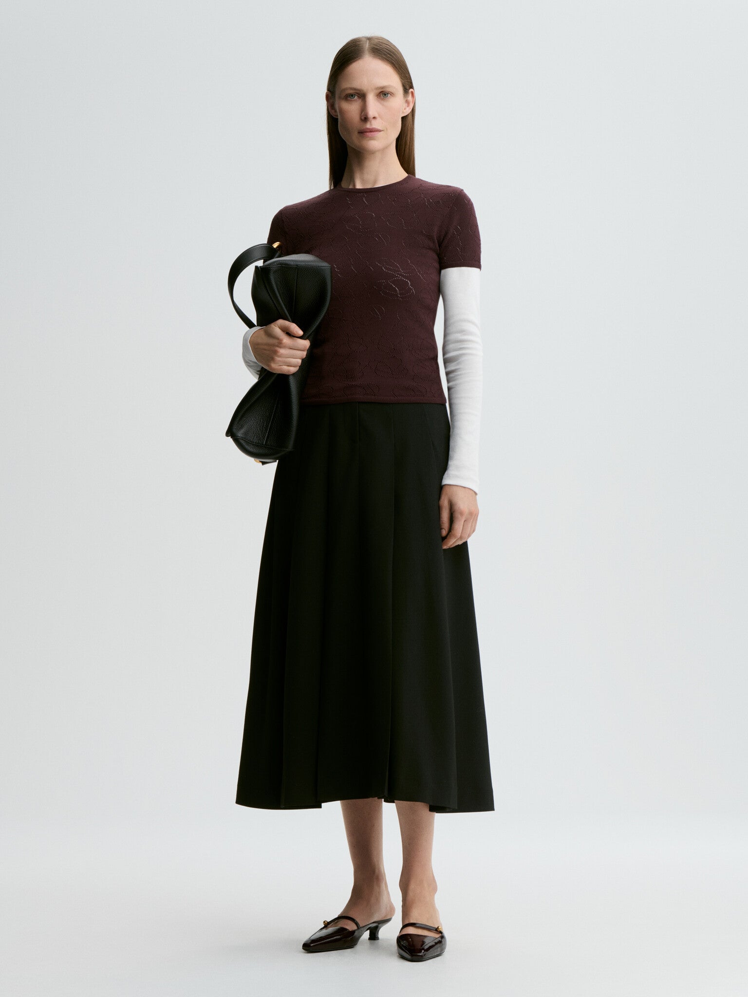 Woman wearing a maroon sweater and black skirt holding a black bag on a white background