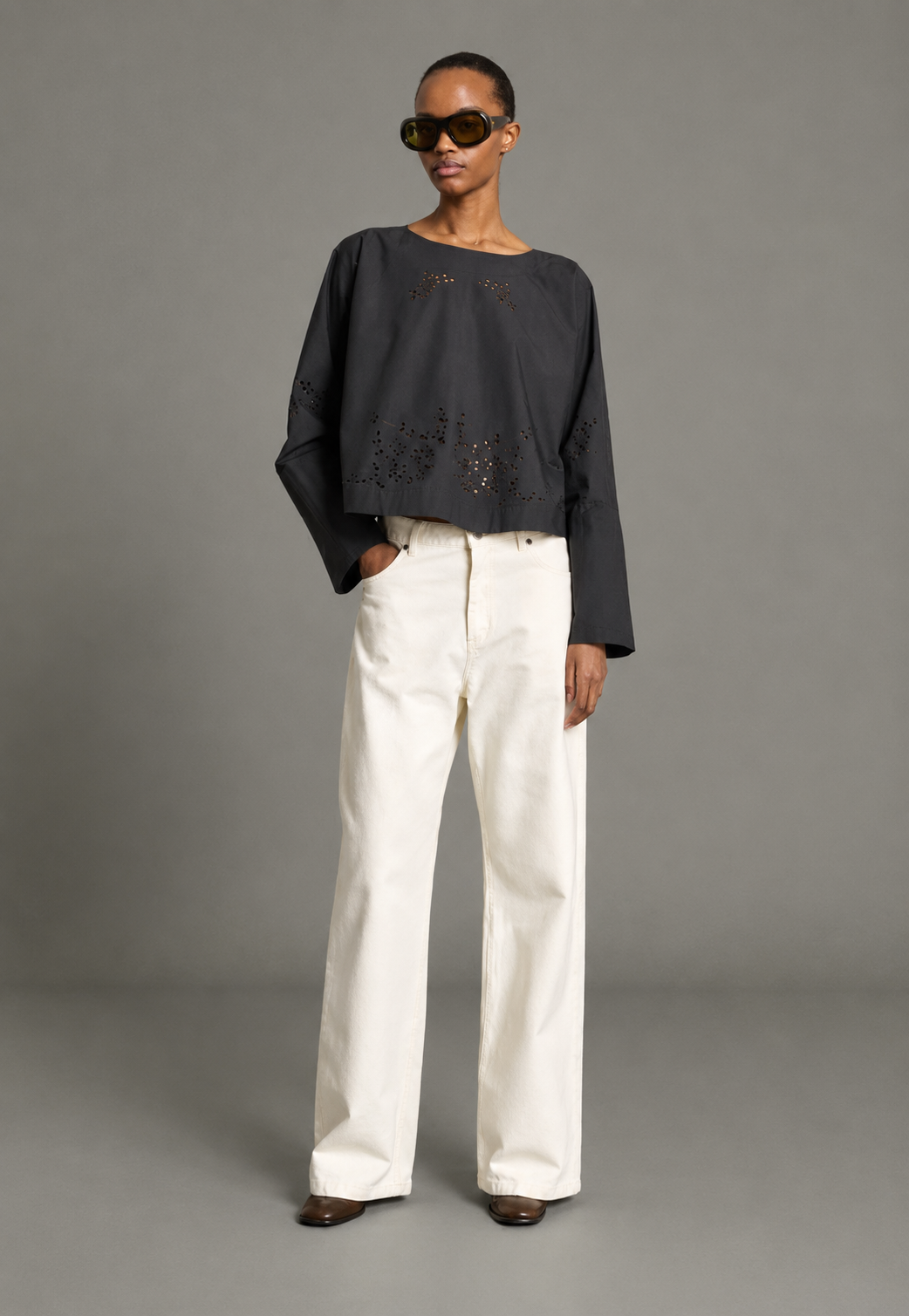 A model stands against a neutral gray studio background wearing a dark charcoal gray long-sleeve top with laser-cutout details and white wide-leg trousers.