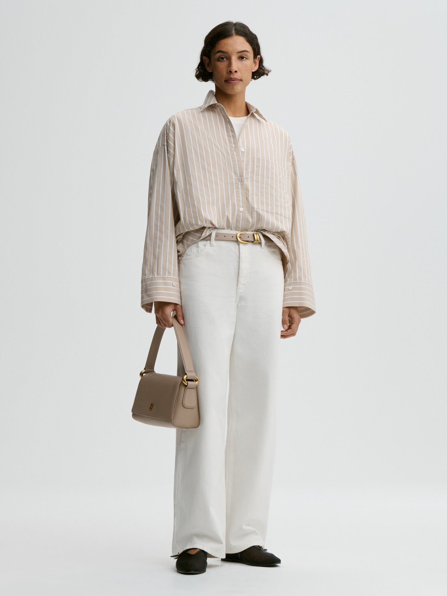 Woman wearing a beige striped shirt and white pants, holding a beige handbag on a light gray background