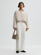 Woman wearing a beige striped shirt and white pants, holding a beige handbag on a light gray background