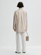 Person wearing a beige striped shirt and white pants, holding a beige handbag on a light gray background