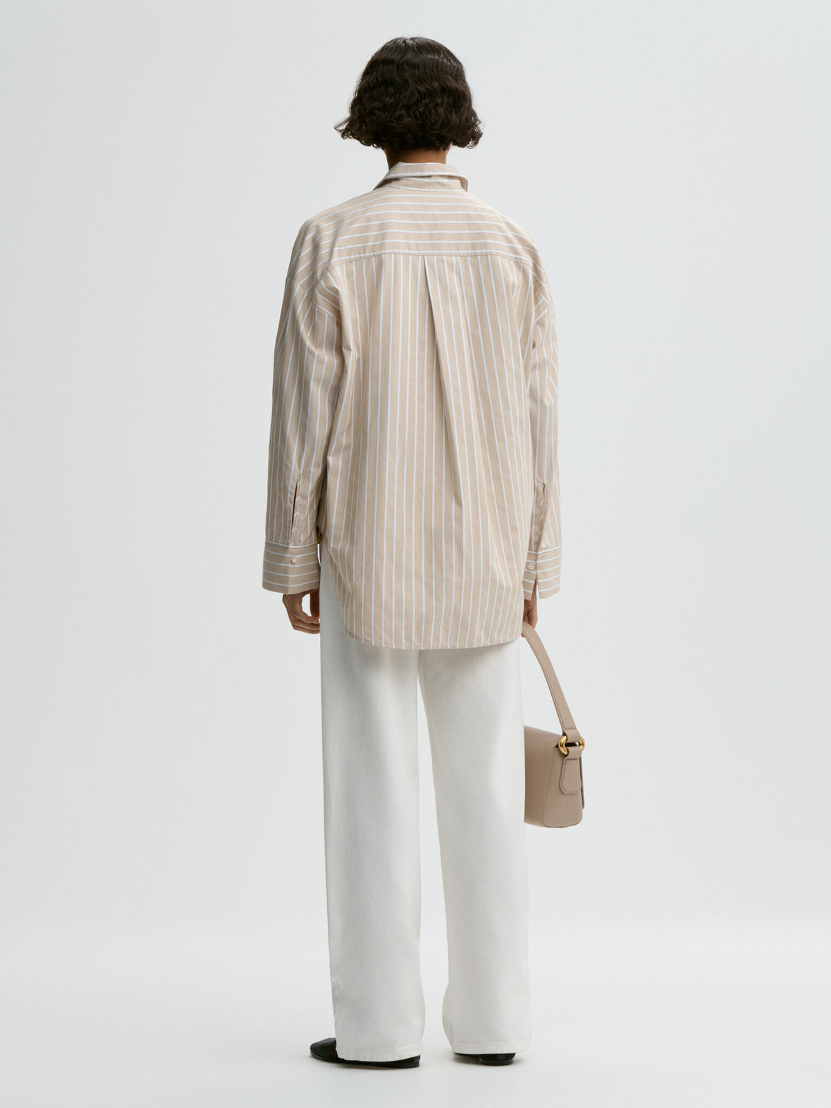Person wearing a beige striped shirt and white pants, holding a beige handbag on a light gray background