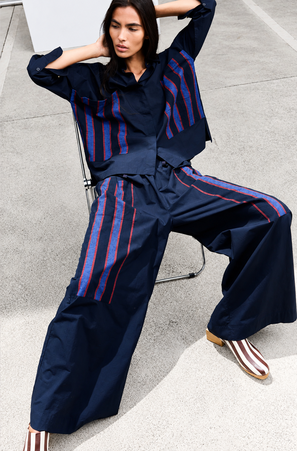 A model wearing a matching navy blue two-piece outfit with red and blue striped details, paired with striped loafers, sitting on a metal chair on a concrete surface.