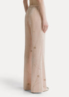 A model shown from the back wearing high-waisted, wide-leg trousers in a light blush pink fabric with subtle gold floral embroidery, set against a plain light gray studio background.