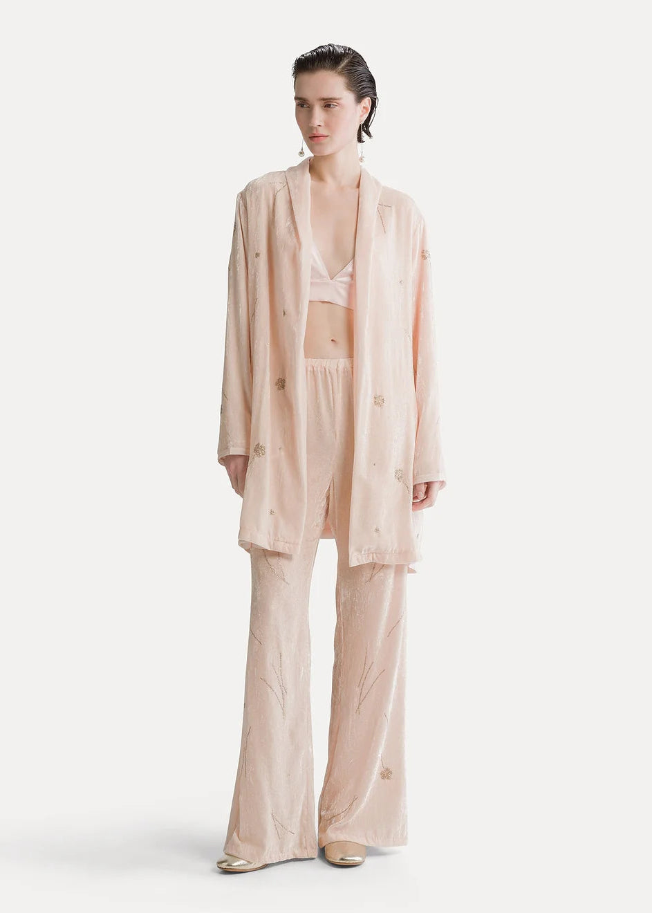 A model stands against a plain off-white studio background wearing a light blush pink matching lounge set consisting of a long open-front robe, a bralette, and wide-leg pants. The fabric has a subtle textured, slightly sheer quality with small tonal embroidered details.