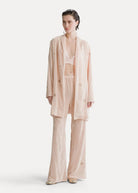 A model stands against a plain off-white studio background wearing a light blush pink matching lounge set consisting of a long open-front robe, a bralette, and wide-leg pants. The fabric has a subtle textured, slightly sheer quality with small tonal embroidered details.