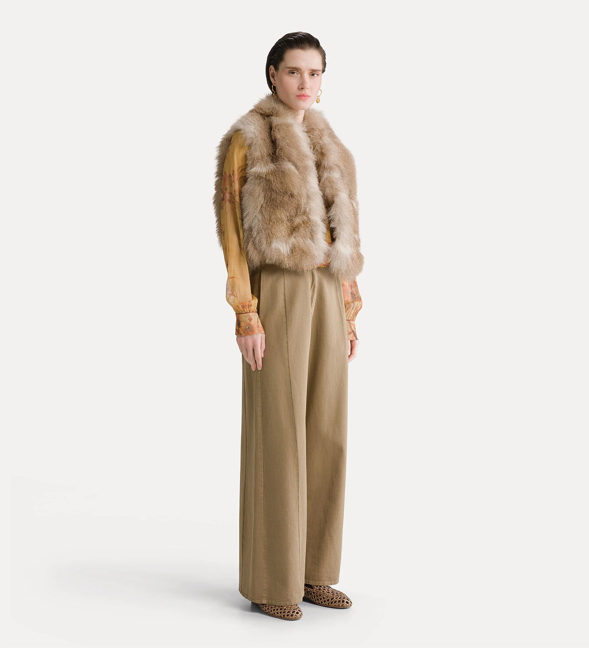 A model stands against a light gray studio background wearing a tan faux-fur vest over a mustard-yellow long-sleeve top, paired with wide-leg beige trousers and brown shoes.