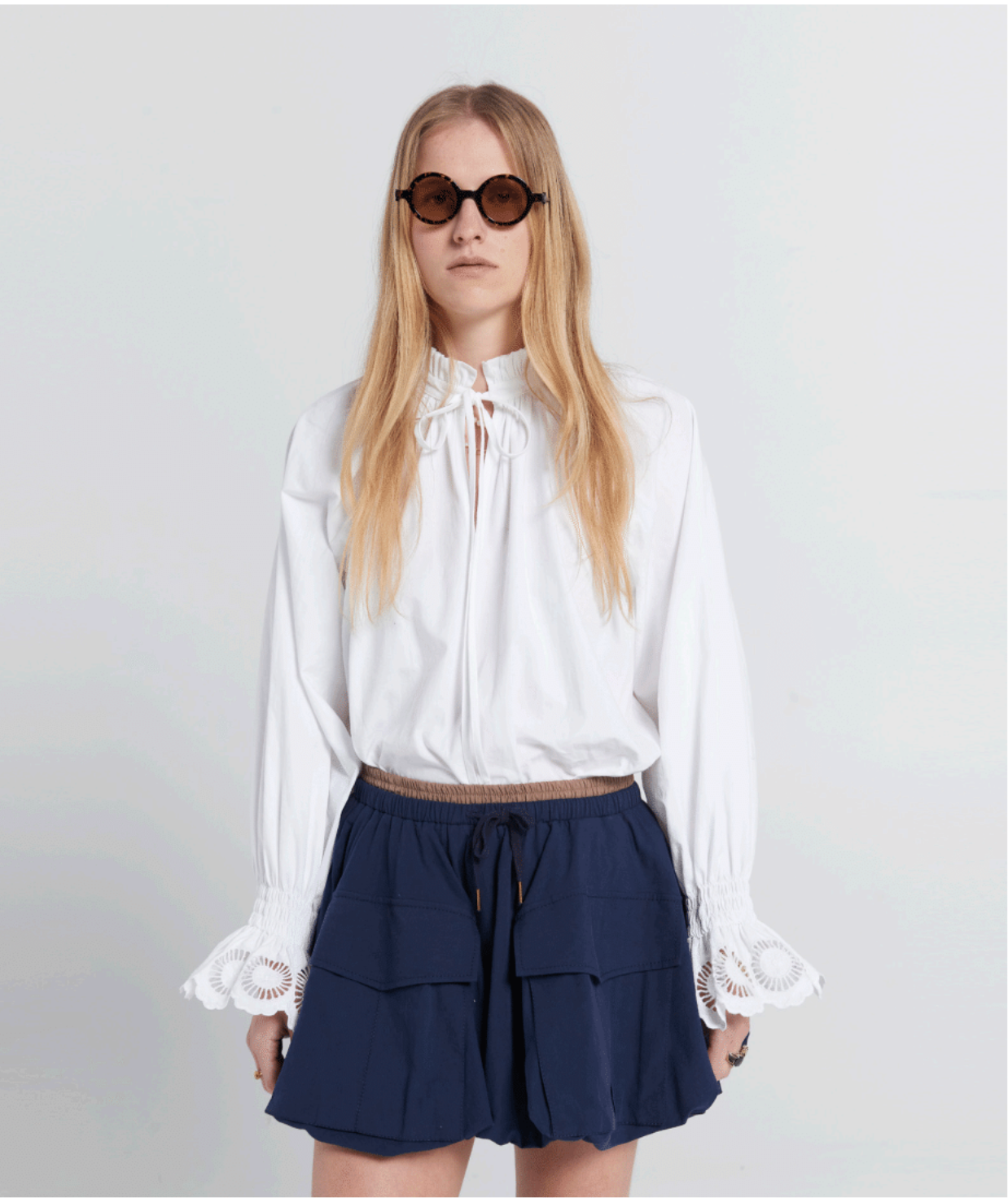 Woman wearing a white blouse and navy skirt with sunglasses on a plain background