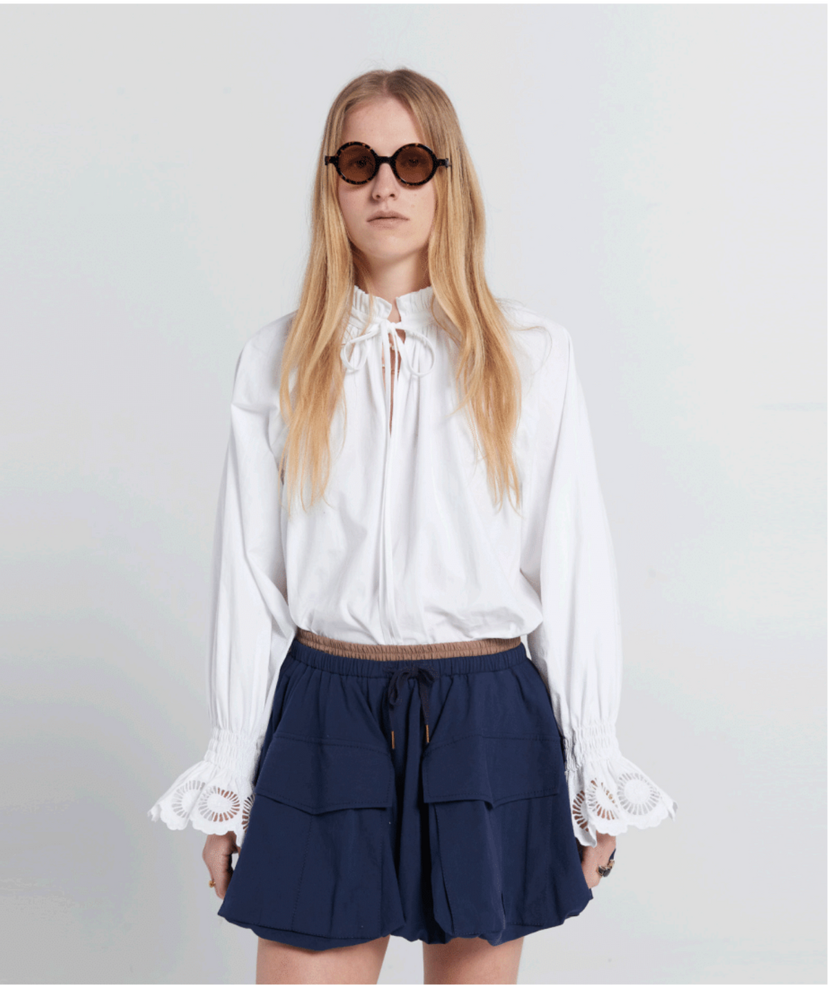 Woman wearing a white blouse and navy skirt with sunglasses on a plain background