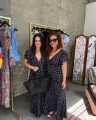 Two women in matching black and white polka dot dresses standing in a room with clothing racks and a screen in the background.