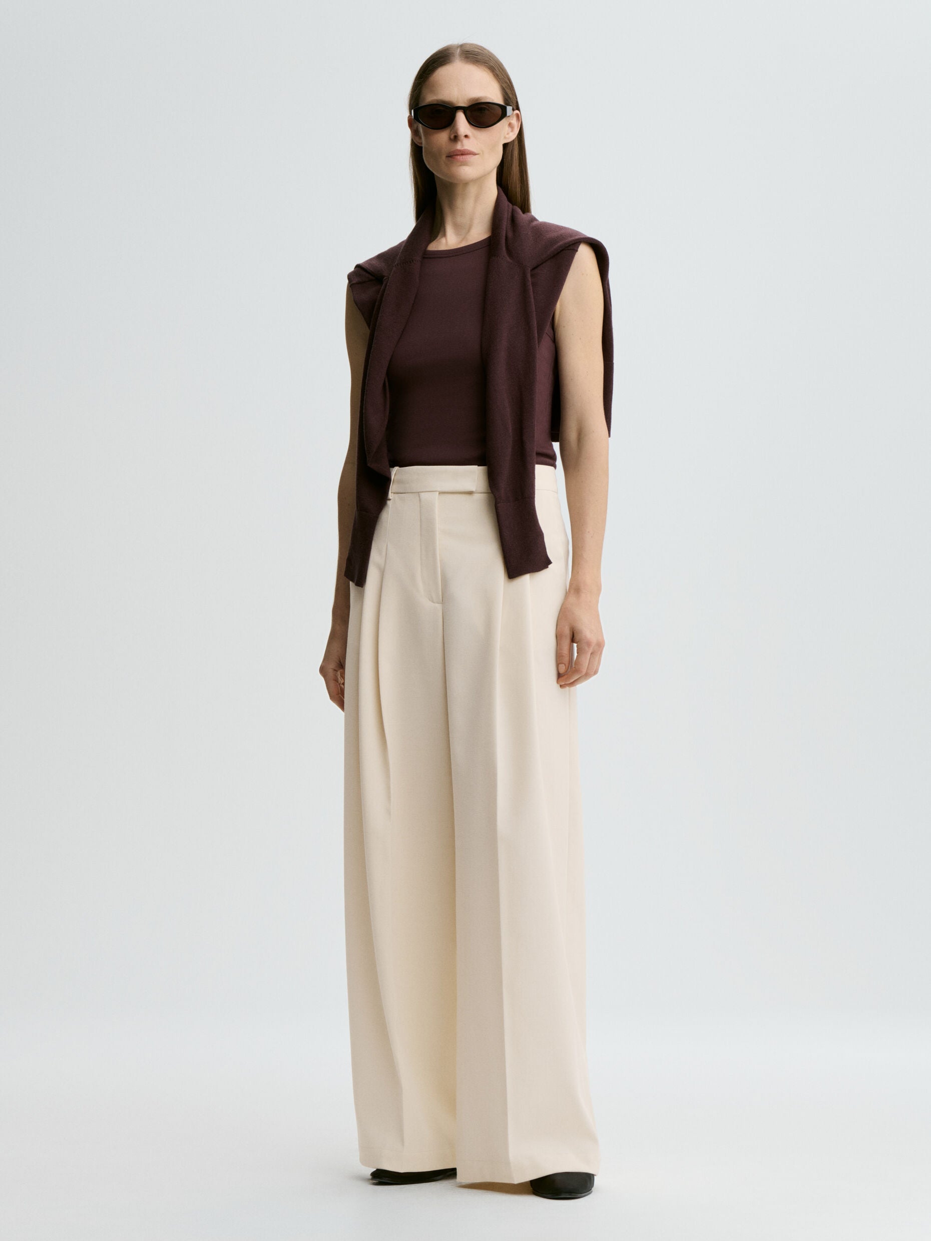 Woman wearing a brown top, beige wide-leg pants, and sunglasses on a light gray background